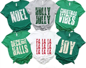 May include: Six short-sleeved crew neck t-shirts in green, gray, and white. The shirts feature distressed-style text prints: NOEL, HOLLY JOLLY, CHRISTMAS VIBES, DECK THE HALLS, FA LA LA LA LA LA LA LA LA, and JOY in white, green, or red ink.