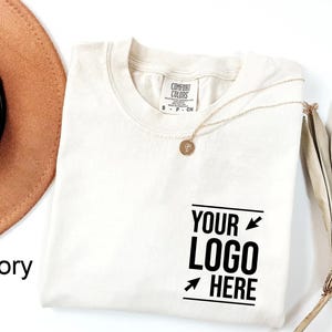 May include: An ivory-colored crew neck t-shirt with a Comfort Colors label at the inner neckline. The front features black text that reads YOUR LOGO HERE, framed by two small arrows pointing toward the center of the text block.