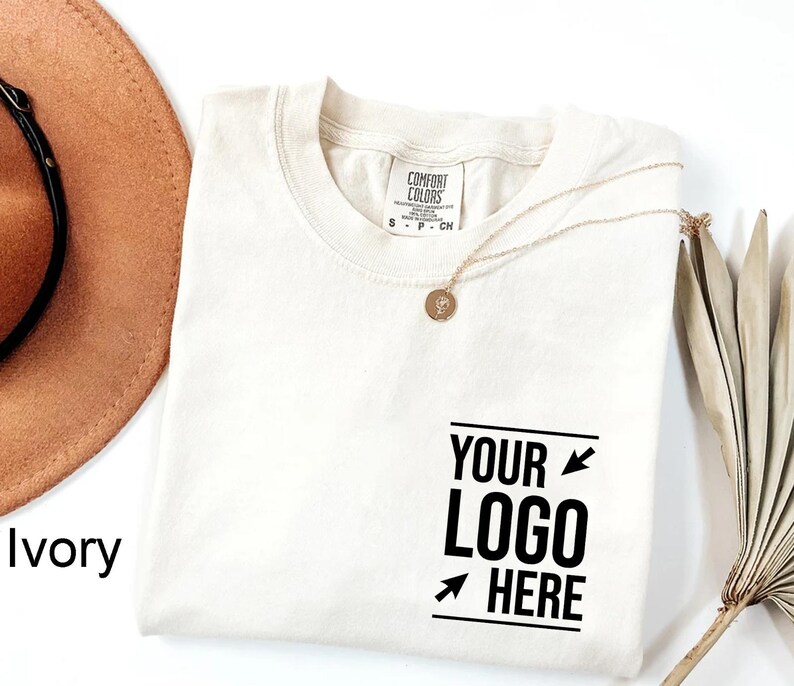 May include: An ivory-colored crew neck t-shirt with a Comfort Colors label at the inner neckline. The front features black text that reads YOUR LOGO HERE, framed by two small arrows pointing toward the center of the text block.