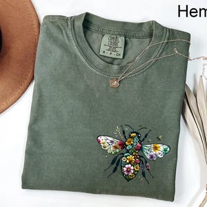 May include: Olive green t-shirt with a floral bee graphic. The bee has colorful wings and is surrounded by flowers. The shirt has a crew neck and short sleeves. A gold necklace with a small pendant is on the shirt. The word "Hemp" is in the top right corner.