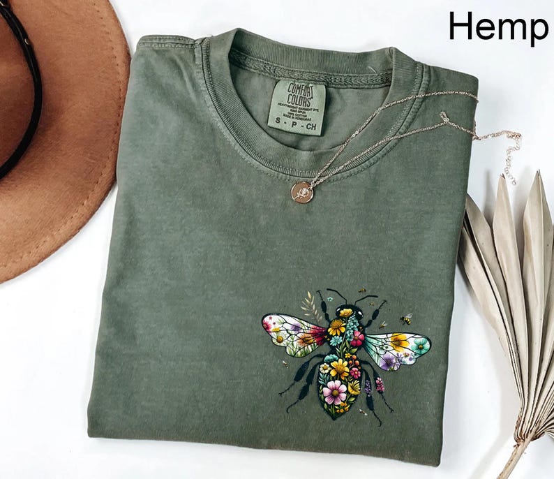 May include: Olive green t-shirt with a floral bee graphic. The bee has colorful wings and is surrounded by flowers. The shirt has a crew neck and short sleeves. A gold necklace with a small pendant is on the shirt. The word "Hemp" is in the top right corner.