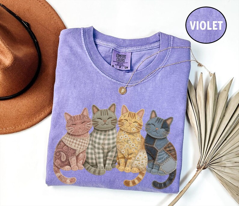 May include: A violet Comfort Colors t-shirt with a graphic of four smiling cats in various patterns and colours. The cats are in shades of pink, grey, yellow, and blue. A gold necklace and a brown hat are also visible.