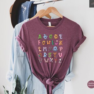 May include: Heather maroon t-shirt featuring the alphabet, with each letter designed as a different animal. The shirt is hanging on a wooden hanger. The shirt has the words "Heather Maroon" printed on the bottom right.
