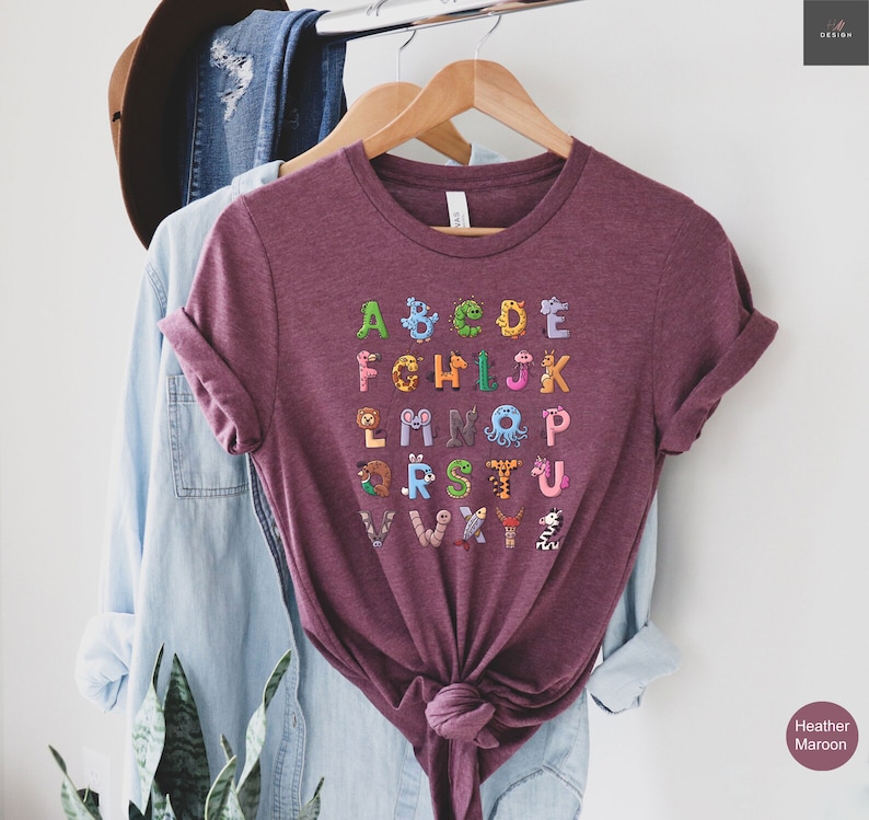 May include: Heather maroon t-shirt featuring the alphabet, with each letter designed as a different animal. The shirt is hanging on a wooden hanger. The shirt has the words "Heather Maroon" printed on the bottom right.