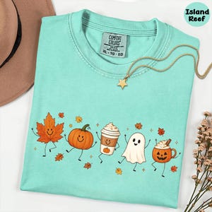K&ouml;nnte beinhalten: Ein mintgr&uuml;nes T-Shirt mit einem Grafikdruck aus einem l&auml;chelnden Ahornblatt, einem K&uuml;rbis, einem Kaffeebecher, einem Geist und einer K&uuml;rbistasse. Das T-Shirt hat ein Comfort Colors-Etikett im Nacken und wird mit einer goldenen Sternkette kombiniert.