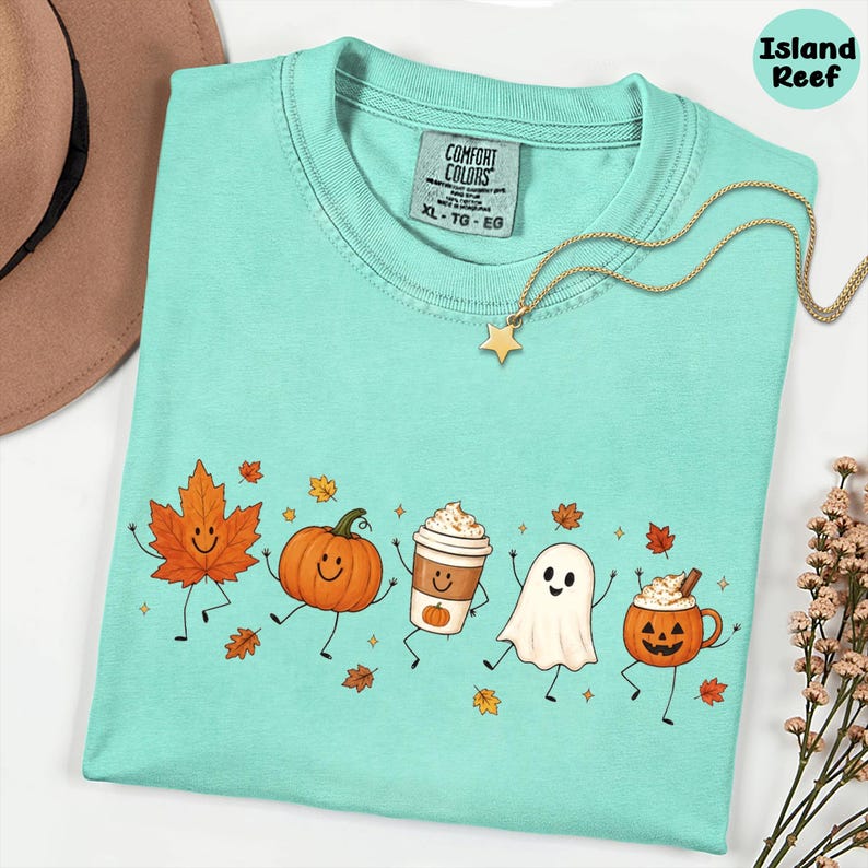 K&ouml;nnte beinhalten: Ein mintgr&uuml;nes T-Shirt mit einem Grafikdruck aus einem l&auml;chelnden Ahornblatt, einem K&uuml;rbis, einem Kaffeebecher, einem Geist und einer K&uuml;rbistasse. Das T-Shirt hat ein Comfort Colors-Etikett im Nacken und wird mit einer goldenen Sternkette kombiniert.