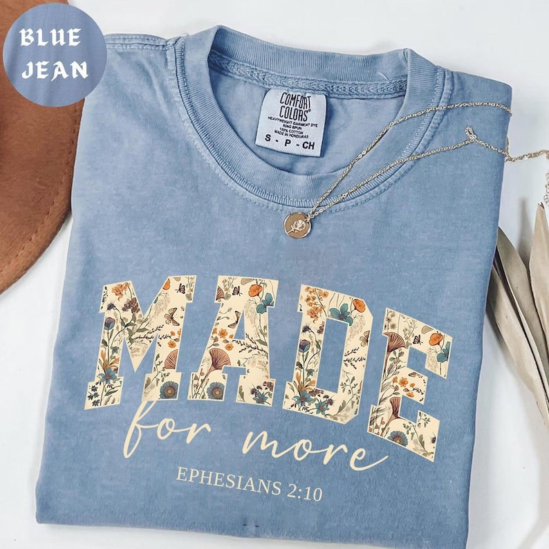 May include: Blue jean Comfort Colors t-shirt with the words "MADE for more" in a floral design. The text "Ephesians 2:10" is printed below. A gold necklace with a pendant is on the shirt.