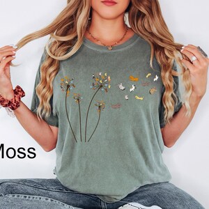 May include: A moss green short-sleeve t-shirt featuring a graphic print of three dandelion stalks. The dandelion heads are composed of small, multi-coloured cats that appear to be blowing away into the air as individual cat figures.