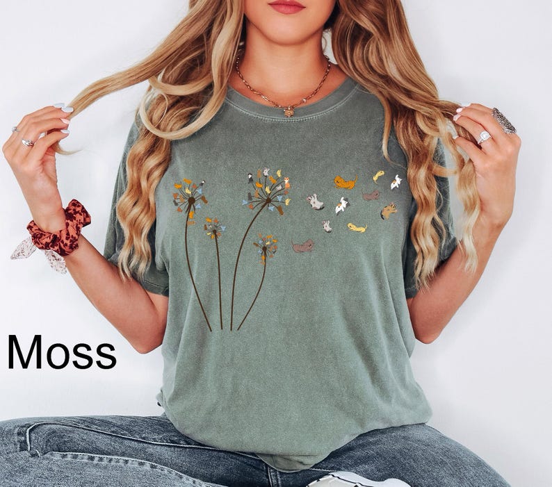 May include: A moss green short-sleeve t-shirt featuring a graphic print of three dandelion stalks. The dandelion heads are composed of small, multi-coloured cats that appear to be blowing away into the air as individual cat figures.