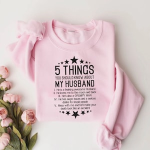 May include: A light pink sweatshirt with the text "5 THINGS YOU SHOULD KNOW ABOUT MY HUSBAND" in black. The text is surrounded by stars. The sweatshirt has long sleeves and a crew neck.