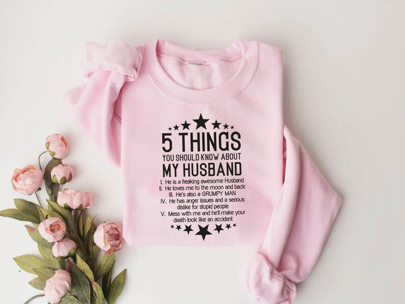 May include: A light pink sweatshirt with the text "5 THINGS YOU SHOULD KNOW ABOUT MY HUSBAND" in black. The text is surrounded by stars. The sweatshirt has long sleeves and a crew neck.