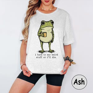 May include: A light grey t-shirt featuring a graphic illustration of a green frog holding a small yellow mug. Below the frog, the text reads: i have to say weird stuff or i'll die. The shirt has short sleeves and a crew neckline.