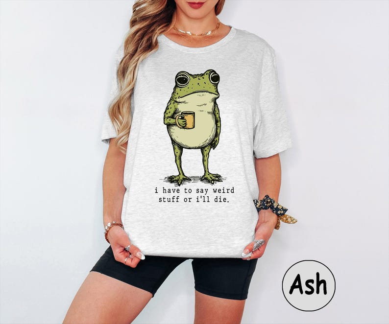 May include: A light grey t-shirt featuring a graphic illustration of a green frog holding a small yellow mug. Below the frog, the text reads: i have to say weird stuff or i'll die. The shirt has short sleeves and a crew neckline.