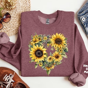 Pode incluir: Camisola de cor bord&ocirc; escuro mesclado com um design floral em forma de cora&ccedil;&atilde;o de girass&oacute;is amarelos e vegeta&ccedil;&atilde;o. A camisola tem gola redonda e mangas compridas. A imagem tamb&eacute;m inclui &oacute;culos de sol, sapatos e cal&ccedil;&otilde;es de ganga.