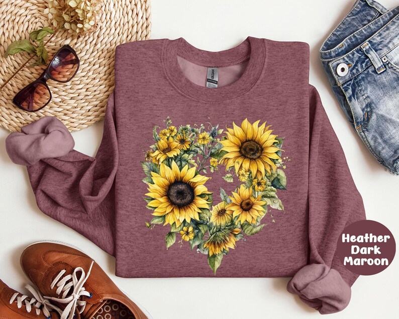 Pode incluir: Camisola de cor bord&ocirc; escuro mesclado com um design floral em forma de cora&ccedil;&atilde;o de girass&oacute;is amarelos e vegeta&ccedil;&atilde;o. A camisola tem gola redonda e mangas compridas. A imagem tamb&eacute;m inclui &oacute;culos de sol, sapatos e cal&ccedil;&otilde;es de ganga.