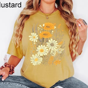 K&ouml;nnte beinhalten: Senffarbenes T-Shirt mit einem Blumendesign aus gelben und wei&szlig;en G&auml;nsebl&uuml;mchen. Das Shirt hat kurze &Auml;rmel und eine lockere Passform. Das Wort "Mustard" ist in Schwarz in der oberen linken Ecke aufgedruckt.