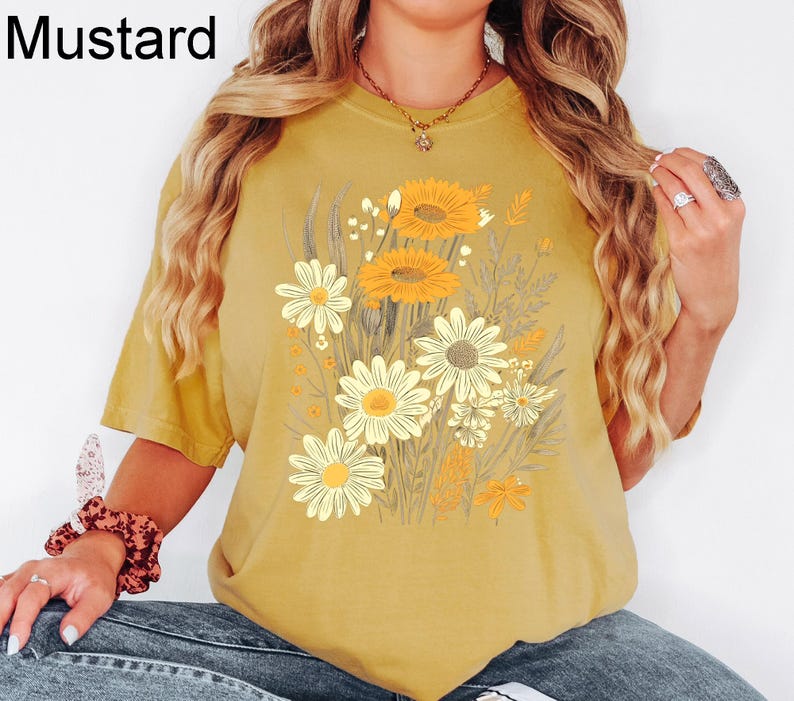 K&ouml;nnte beinhalten: Senffarbenes T-Shirt mit einem Blumendesign aus gelben und wei&szlig;en G&auml;nsebl&uuml;mchen. Das Shirt hat kurze &Auml;rmel und eine lockere Passform. Das Wort "Mustard" ist in Schwarz in der oberen linken Ecke aufgedruckt.