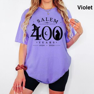 Puede incluir: Una camiseta violeta de manga corta con un estampado gr&aacute;fico negro que dice SALEM MASSACHUSETTS 400 YEARS 1626-2026, acompa&ntilde;ado de ilustraciones de un sombrero de bruja, una luna creciente y la silueta de un gato en el pecho.