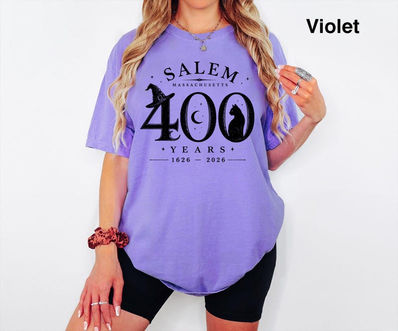 Puede incluir: Una camiseta violeta de manga corta con un estampado gr&aacute;fico negro que dice SALEM MASSACHUSETTS 400 YEARS 1626-2026, acompa&ntilde;ado de ilustraciones de un sombrero de bruja, una luna creciente y la silueta de un gato en el pecho.