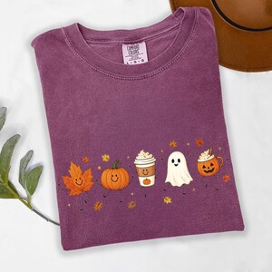 K&ouml;nnte beinhalten: Ein pflaumenfarbenes T-Shirt mit Rundhalsausschnitt und einem Grafikdruck, der ein l&auml;chelndes orangefarbenes Ahornblatt, einen K&uuml;rbis, eine Kaffeetasse, ein wei&szlig;es Gespenst und eine Halloween-Laterne zeigt, verziert mit kleinen Herbstbl&auml;ttern und Sternen.