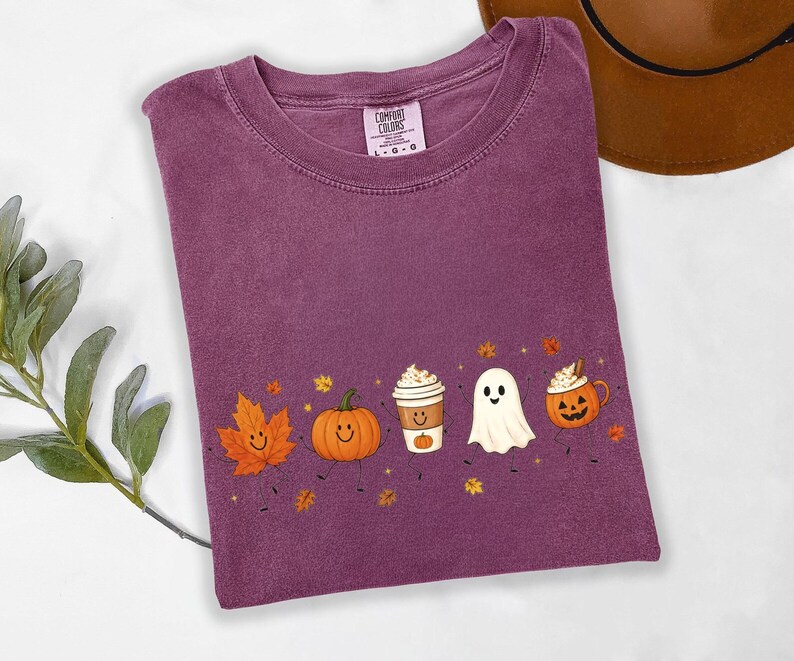 K&ouml;nnte beinhalten: Ein pflaumenfarbenes T-Shirt mit Rundhalsausschnitt und einem Grafikdruck, der ein l&auml;chelndes orangefarbenes Ahornblatt, einen K&uuml;rbis, eine Kaffeetasse, ein wei&szlig;es Gespenst und eine Halloween-Laterne zeigt, verziert mit kleinen Herbstbl&auml;ttern und Sternen.
