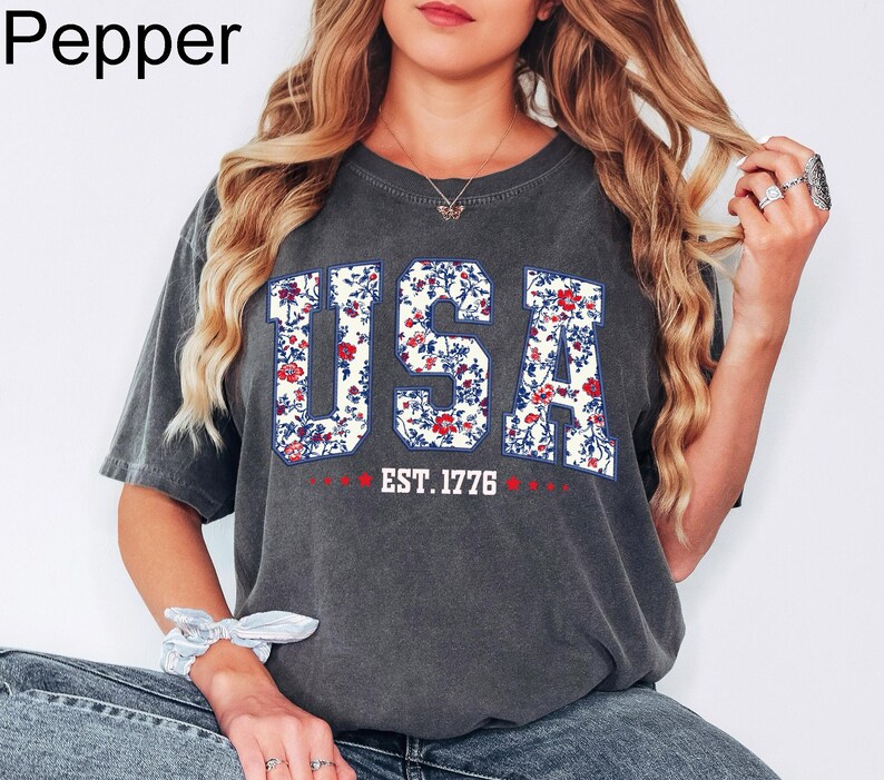 May include: A dark gray t-shirt with the word "USA" in a floral design. The letters are filled with red and blue flowers. Below the word "USA" is the text "EST. 1776" in red.