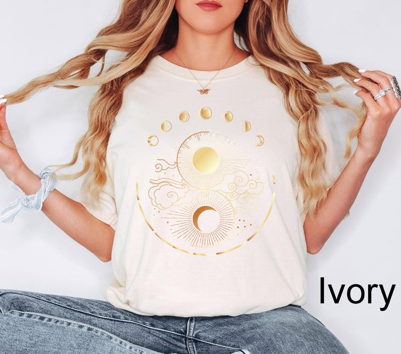 Pu&ograve; includere: T-shirt color avorio con un design celeste dorato con illustrazioni di sole, luna e fasi. La maglietta ha scollo tondo e maniche corte. Il testo "Ivory" &egrave; visualizzato in nero in basso a destra.