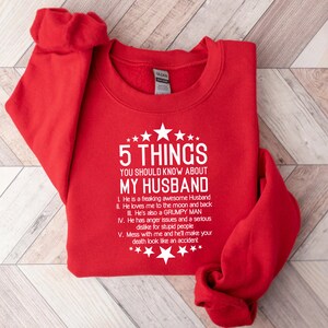 May include: A red sweatshirt with white text that reads "5 THINGS YOU SHOULD KNOW ABOUT MY HUSBAND." The text is followed by a list of five humorous statements about a husband. The sweatshirt has a crew neck and long sleeves.