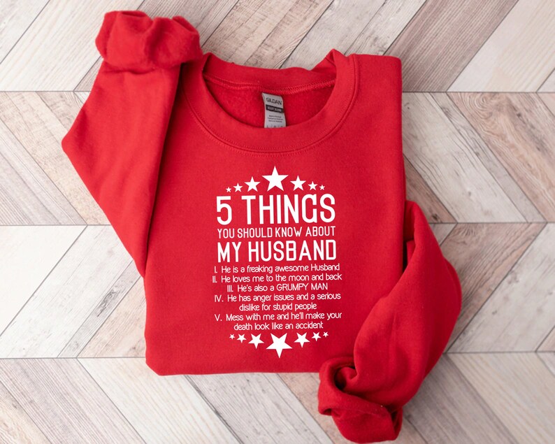May include: A red sweatshirt with white text that reads "5 THINGS YOU SHOULD KNOW ABOUT MY HUSBAND." The text is followed by a list of five humorous statements about a husband. The sweatshirt has a crew neck and long sleeves.