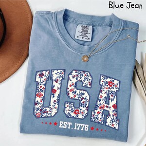 May include: Blue jean-colored t-shirt with the word "USA" in a floral pattern. The shirt also features the text "EST. 1776" and a necklace with a pendant. A brown hat is visible in the upper left corner.