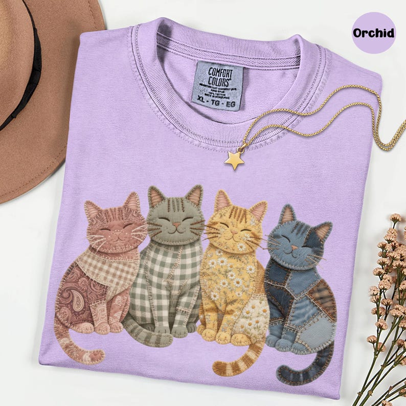 May include: A lilac Comfort Colors t-shirt with four smiling cat illustrations in patchwork designs. A gold star pendant necklace is on the shirt. The shirt is size XL-TG-EG.