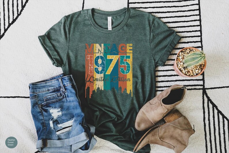 May include: A green t-shirt with the text "VINTAGE 1975 Limited Edition" in a colorful design. The shirt is paired with blue denim shorts, brown shoes, and a small potted cactus.