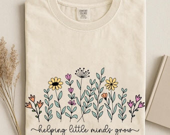 May include: An ivory short-sleeve t-shirt featuring a graphic of colourful wildflowers in yellow, pink, and orange with green stems. Below the flowers, the text reads "helping little minds grow" in a black cursive script font.