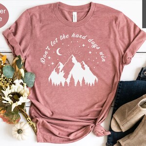 Pu&ograve; includere: T-shirt color malva con grafica bianca di montagne, luna, stelle e la scritta "Don't let the hard days win." La maglietta &egrave; abbinata a jeans blu e sandali beige. Un mazzo di fiori e una piccola zucca sono anche nell'immagine.