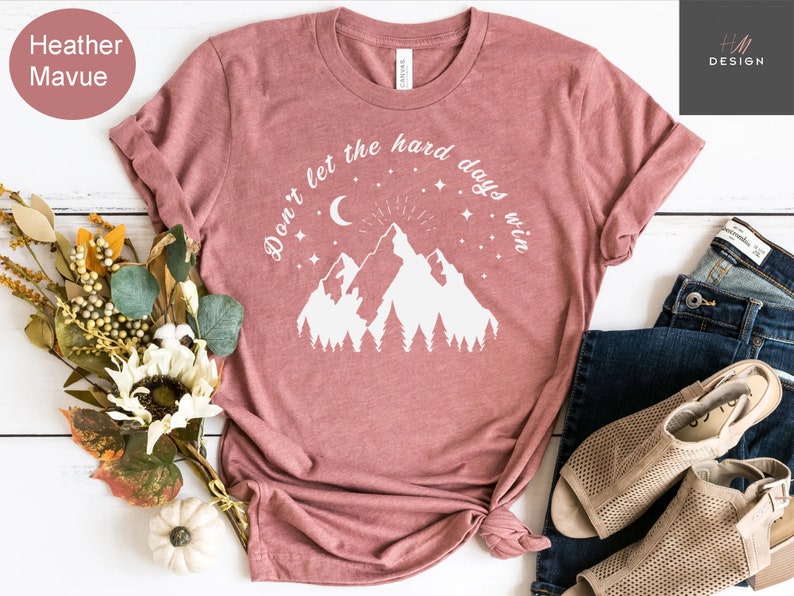 Pu&ograve; includere: T-shirt color malva con grafica bianca di montagne, luna, stelle e la scritta "Don't let the hard days win." La maglietta &egrave; abbinata a jeans blu e sandali beige. Un mazzo di fiori e una piccola zucca sono anche nell'immagine.