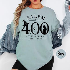 Puede incluir: Una camiseta de cuello redondo azul claro con un gr&aacute;fico negro que dice SALEM MASSACHUSETTS 400 YEARS 1626-2026. El dise&ntilde;o incluye un sombrero de bruja, una luna creciente dentro del cero y la silueta de un gato negro bajo una chaqueta vaquera.