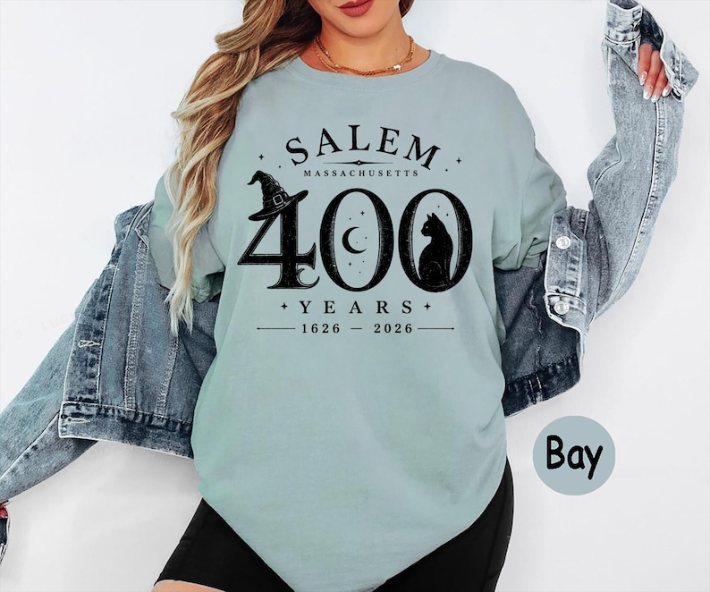 Puede incluir: Una camiseta de cuello redondo azul claro con un gr&aacute;fico negro que dice SALEM MASSACHUSETTS 400 YEARS 1626-2026. El dise&ntilde;o incluye un sombrero de bruja, una luna creciente dentro del cero y la silueta de un gato negro bajo una chaqueta vaquera.