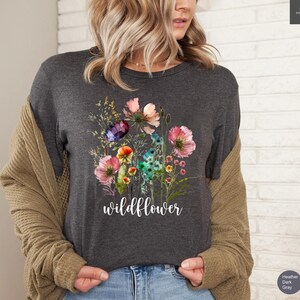 May include: Dark gray t-shirt featuring a watercolor floral design with pink, purple, and blue flowers. The word "wildflower" is written in white script below the flowers. The shirt is a heather dark gray color.