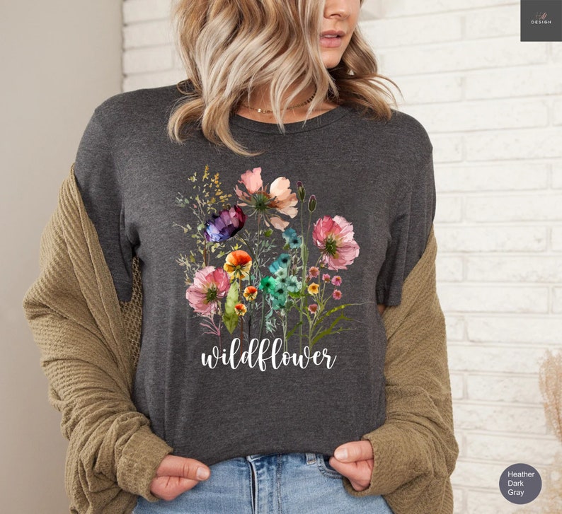 May include: Dark gray t-shirt featuring a watercolor floral design with pink, purple, and blue flowers. The word "wildflower" is written in white script below the flowers. The shirt is a heather dark gray color.