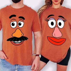 May include: Two rust-colored t-shirts featuring Mr. Potato Head and Mrs. Potato Head faces. The Mr. Potato Head shirt has a black mustache, orange nose, and a smiling mouth. The Mrs. Potato Head shirt has eyelashes, a pink nose, and red lips.