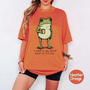 May include: A heather orange t-shirt featuring a graphic of a green frog holding a yellow mug. Below the frog, the text reads: i have to say weird stuff or i'll die. The shirt has short sleeves and a crew neckline, presented in a casual fit.
