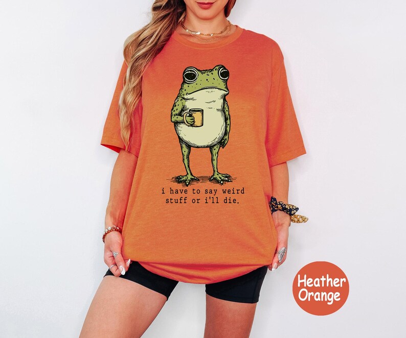May include: A heather orange t-shirt featuring a graphic of a green frog holding a yellow mug. Below the frog, the text reads: i have to say weird stuff or i'll die. The shirt has short sleeves and a crew neckline, presented in a casual fit.