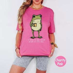 May include: A bright pink t-shirt featuring a green frog illustration holding a small yellow mug. Below the frog, the text reads, "i have to say weird stuff or i'll die." The shirt has a crew neckline and short sleeves, presented in a solid colour.