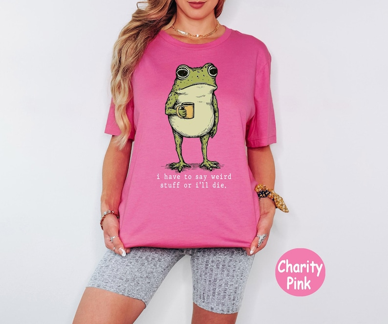 May include: A bright pink t-shirt featuring a green frog illustration holding a small yellow mug. Below the frog, the text reads, "i have to say weird stuff or i'll die." The shirt has a crew neckline and short sleeves, presented in a solid colour.