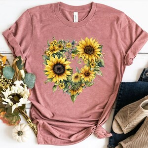 Pode incluir: Uma camiseta rosa empoeirada com um desenho em forma de cora&ccedil;&atilde;o de girass&oacute;is. Os girass&oacute;is t&ecirc;m p&eacute;talas amarelas e centros castanhos escuros, com folhas e caules verdes. A camisa &eacute; combinada com jeans azuis e sand&aacute;lias bege.