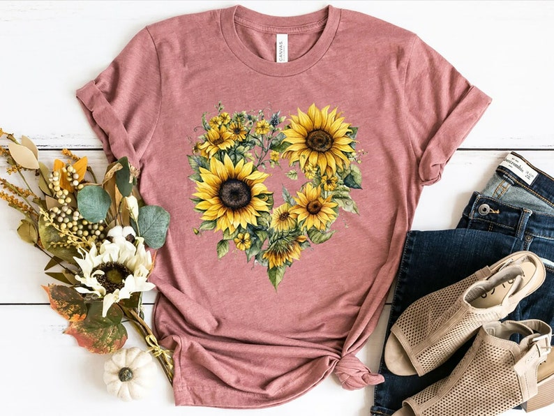 Pode incluir: Uma camiseta rosa empoeirada com um desenho em forma de cora&ccedil;&atilde;o de girass&oacute;is. Os girass&oacute;is t&ecirc;m p&eacute;talas amarelas e centros castanhos escuros, com folhas e caules verdes. A camisa &eacute; combinada com jeans azuis e sand&aacute;lias bege.