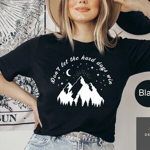 Pu&ograve; includere: T-shirt nera con grafica bianca di montagne, luna crescente, stelle e la scritta "Don't let the hard days win". La maglietta ha anche la parola "Black" in un cerchio nero.