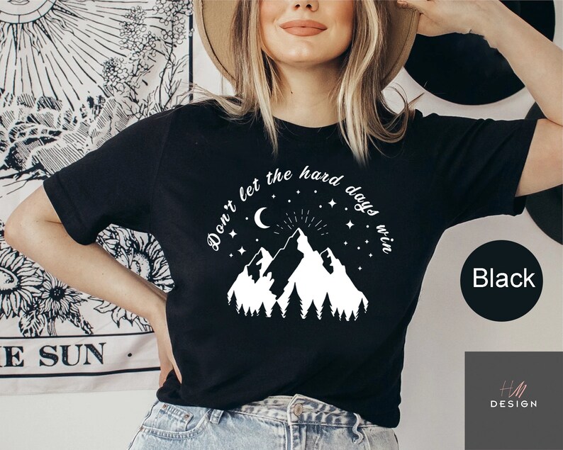 Pu&ograve; includere: T-shirt nera con grafica bianca di montagne, luna crescente, stelle e la scritta "Don't let the hard days win". La maglietta ha anche la parola "Black" in un cerchio nero.