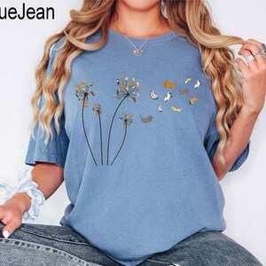 May include: A blue jean coloured short-sleeved t-shirt featuring a graphic print of three dandelion stalks where the seeds are replaced by small, multi-coloured dogs. The text BlueJean is printed in the upper left corner of the image.