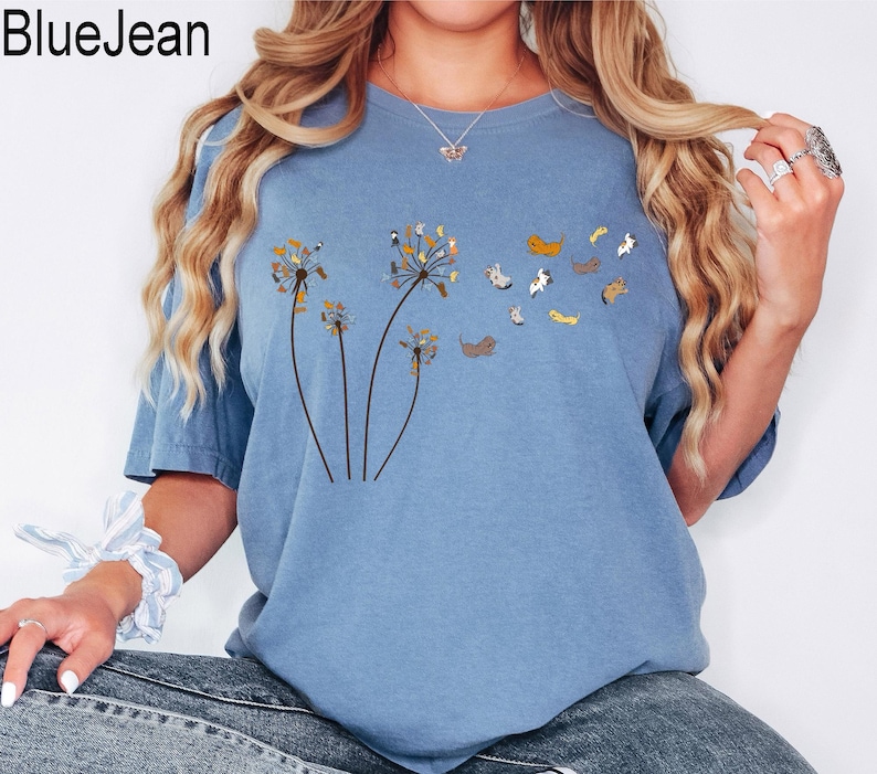 May include: A blue jean coloured short-sleeved t-shirt featuring a graphic print of three dandelion stalks where the seeds are replaced by small, multi-coloured dogs. The text BlueJean is printed in the upper left corner of the image.